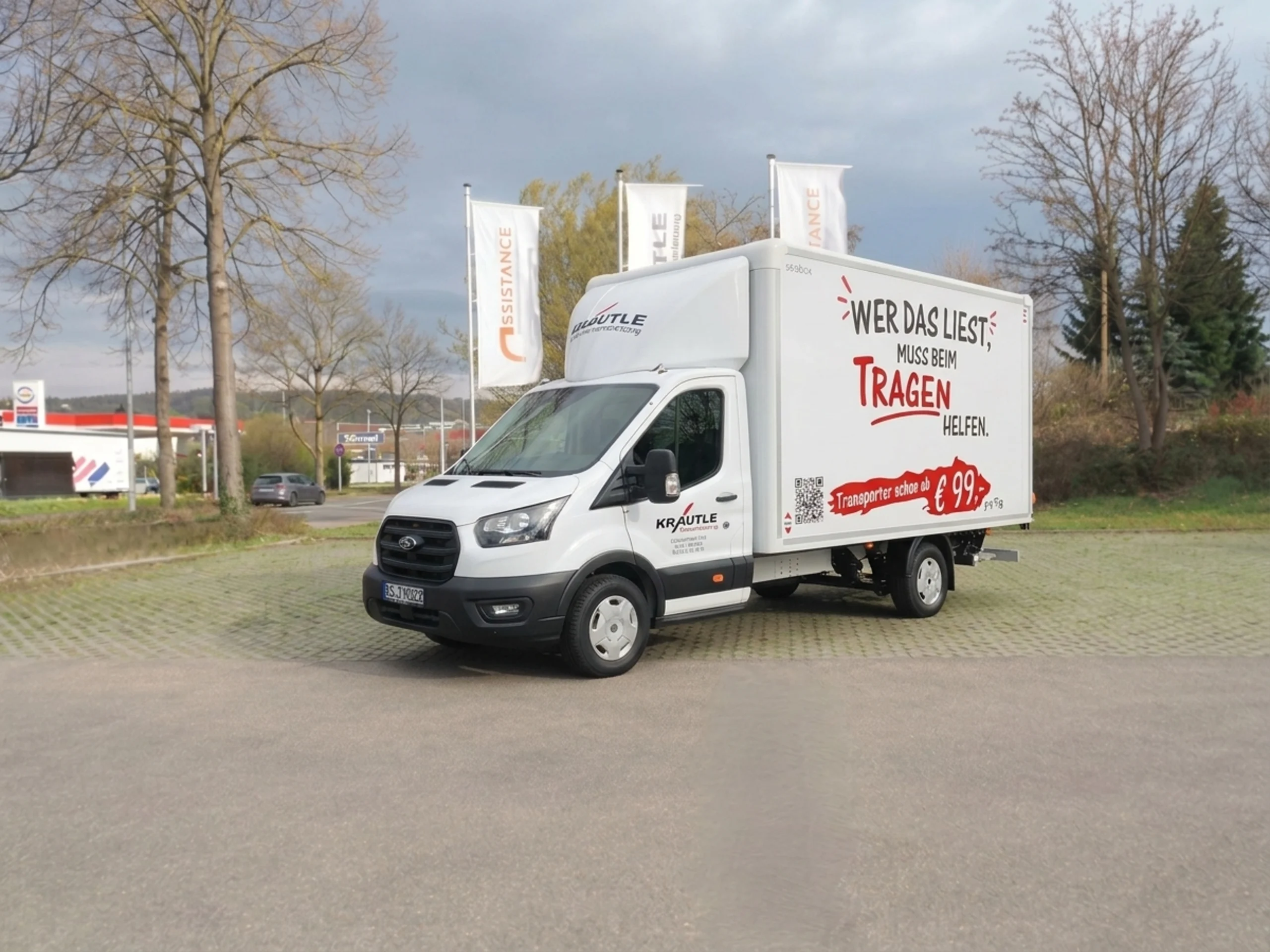 Weißer Transporter mit Werbung: Wer das liest, muss beim Tragen helfen steht auf der Seite, parkend auf einem Platz.